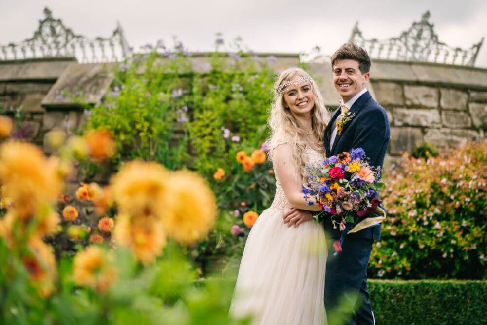 The bride and groom hold each other and smile outside surrounded by flowers.