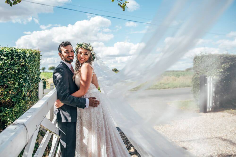 The couple smile and embrace each other outside as the wind sweeps the brides veil.