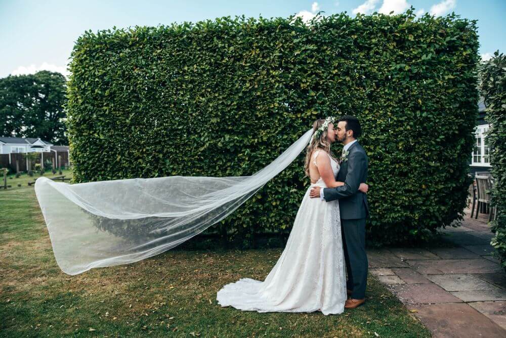 The bride and groom kiss in front of a big hedge.