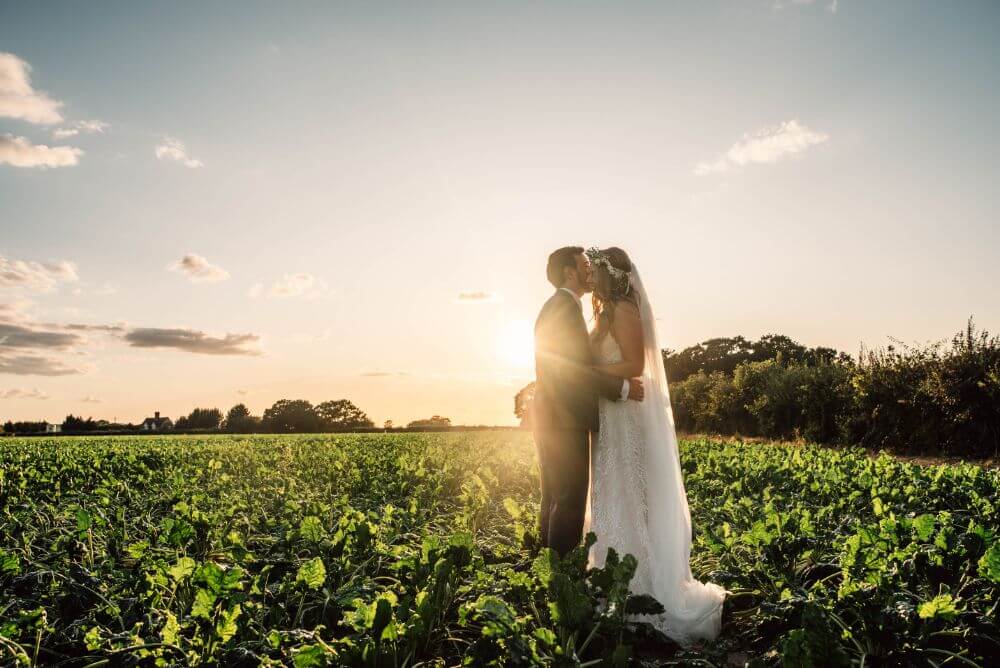 The groom gives the bride a peck on the cheek in the middle of a field.