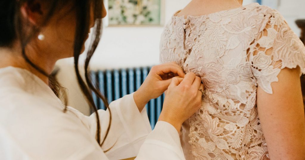 Bride doing up the buttons at the back of lace blush dress of other bride