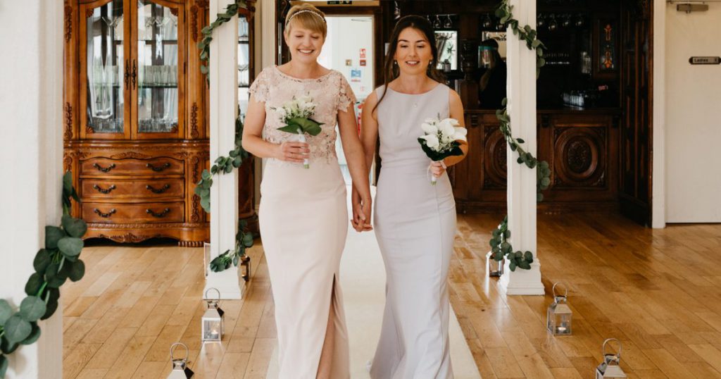 Brides holding hands, walking down aisle holding bouquets of white flowers