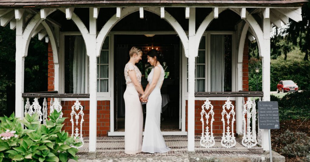 Brides holding hands facing each other outside quaint white and red-brick lodge