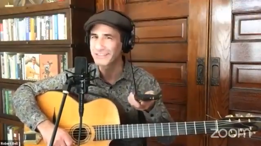 Robert Bell holding a guitar and pointing a remote control at livestream viewers while speaking into a microphone