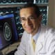 Carlos Pardo-Villamizar wearing a white lab coat in a lab with brain images on screens behind him.