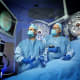 Jena Miller and Ahmet Baschat demonstrate a fetoscope in an OR setting. Both are wearing surgical gowns, caps, masks and gloves and are looking at a monitor screen.