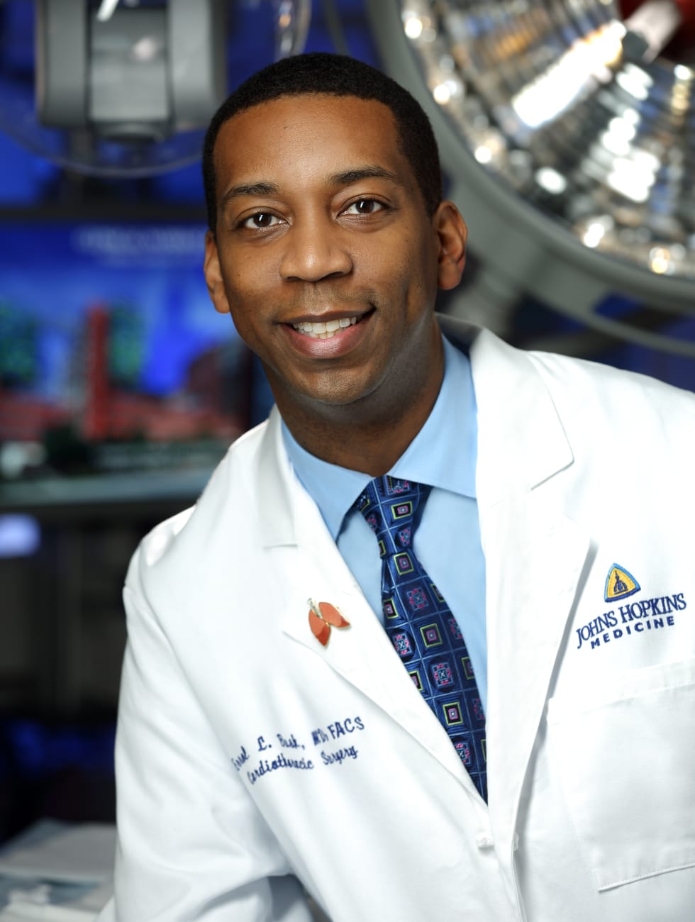 Errol Bush, in a white lab coat, light blue button down shirt and blue tie, poses for a photo in an operating room.