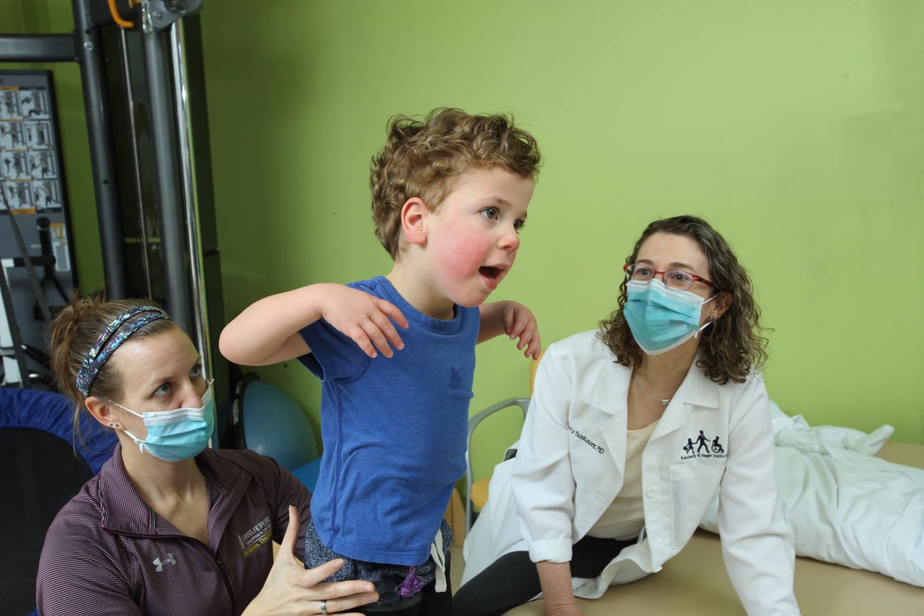 A toddler with curly hair in a blue shirt lifts his arms as he is held by a physical therapist, as Stacy Suskauer look on.