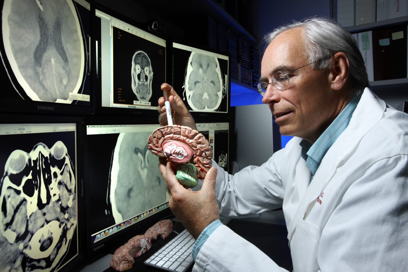 Dr. Dan Hanley holds a plastic brain model while facing computer monitors displaying brain images.