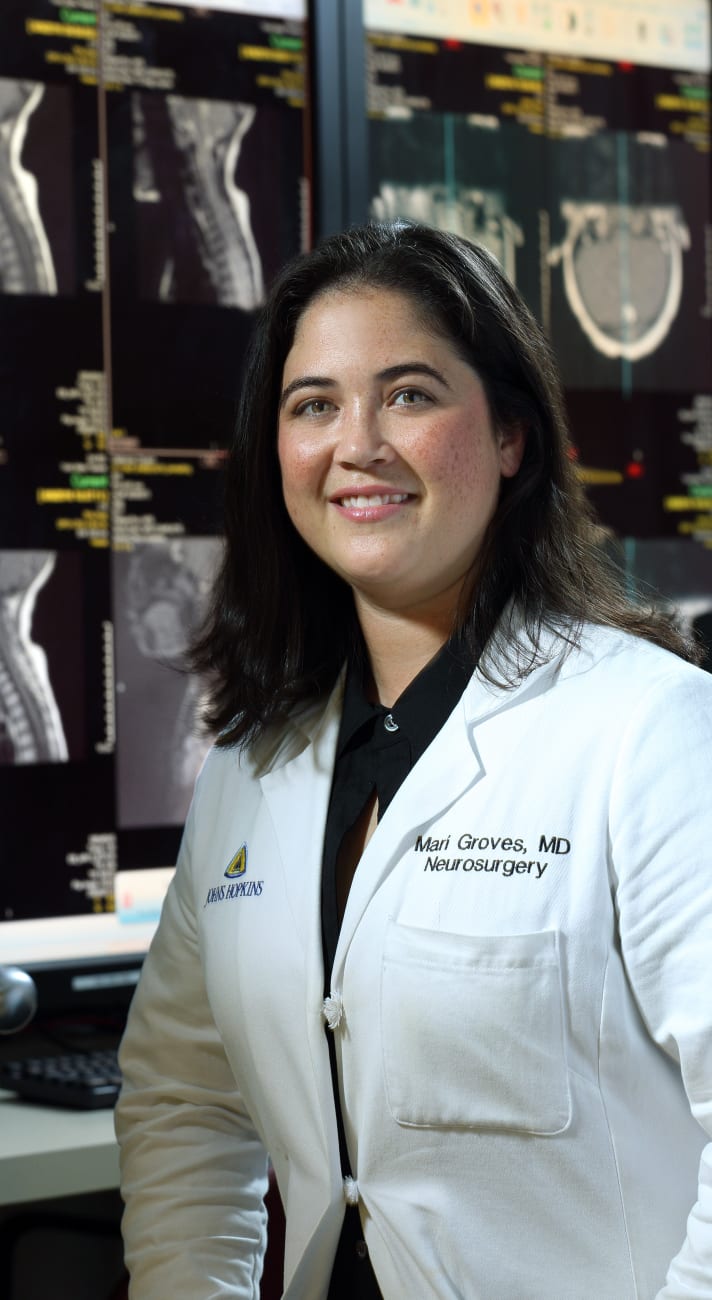 Dr. Mari Groves, pictured in a white lab coat, smiles in a lab with x-rays and other images displayed behind her.