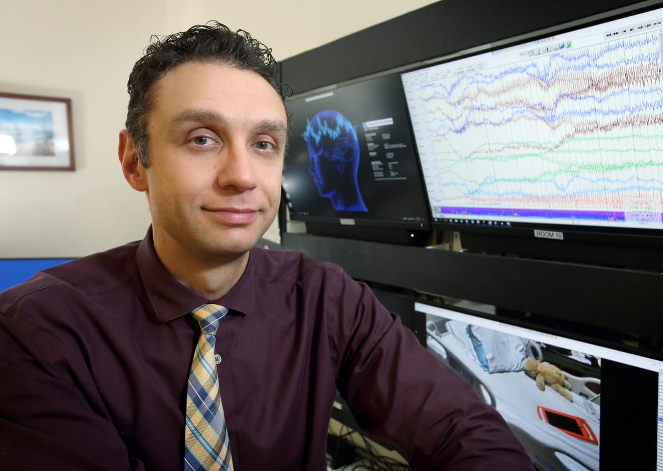 Ahmad Marashly, wearing a purple shirt and plaid tie, poses in front of computer monitors showing brain activity 