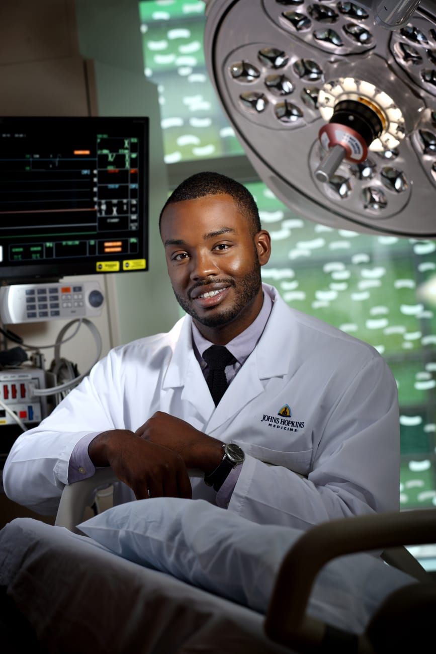 Jason Vaught, wearing a white lab coat, poses in a patient room, leaning on an empty bed, with monitors and a large light behind him.