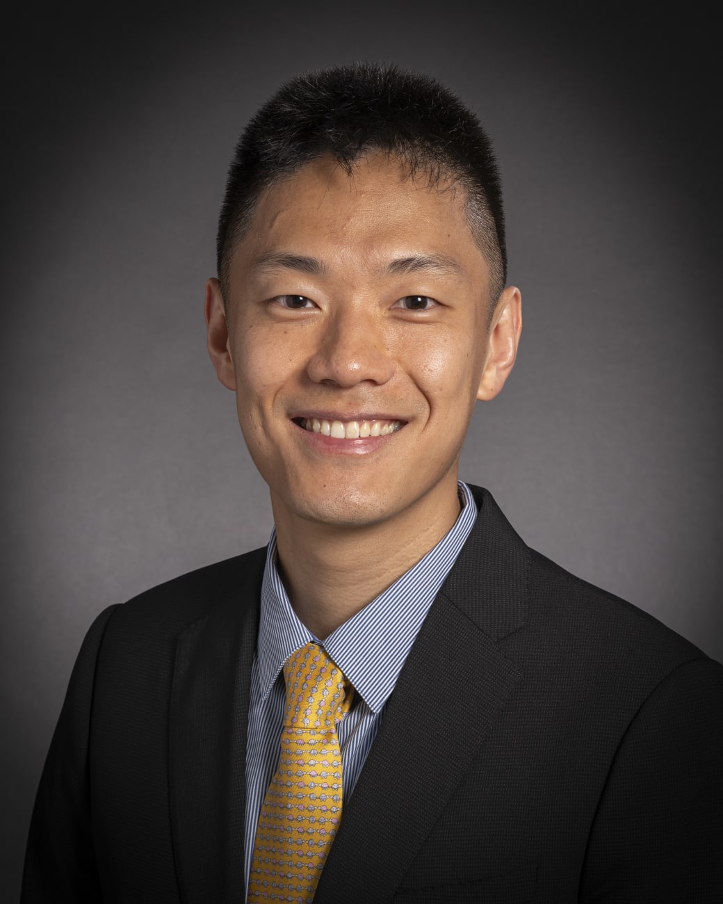 Risheng Xu, in a formal portrait, wearing a black suit and light blue striped button down shirt with a yellow tie