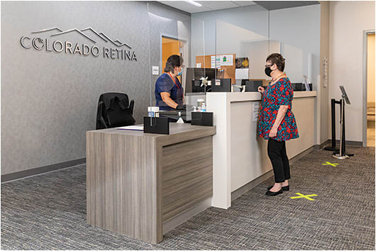 Capitalizing on Lean concepts, the new offices at Colorado Retina were designed without a traditional waiting room. Here Phyllis Romero, front desk representative (left), assists a patient at check-in. When check-in is complete, Ms. Romero electronically moves the patient into the workflow system.