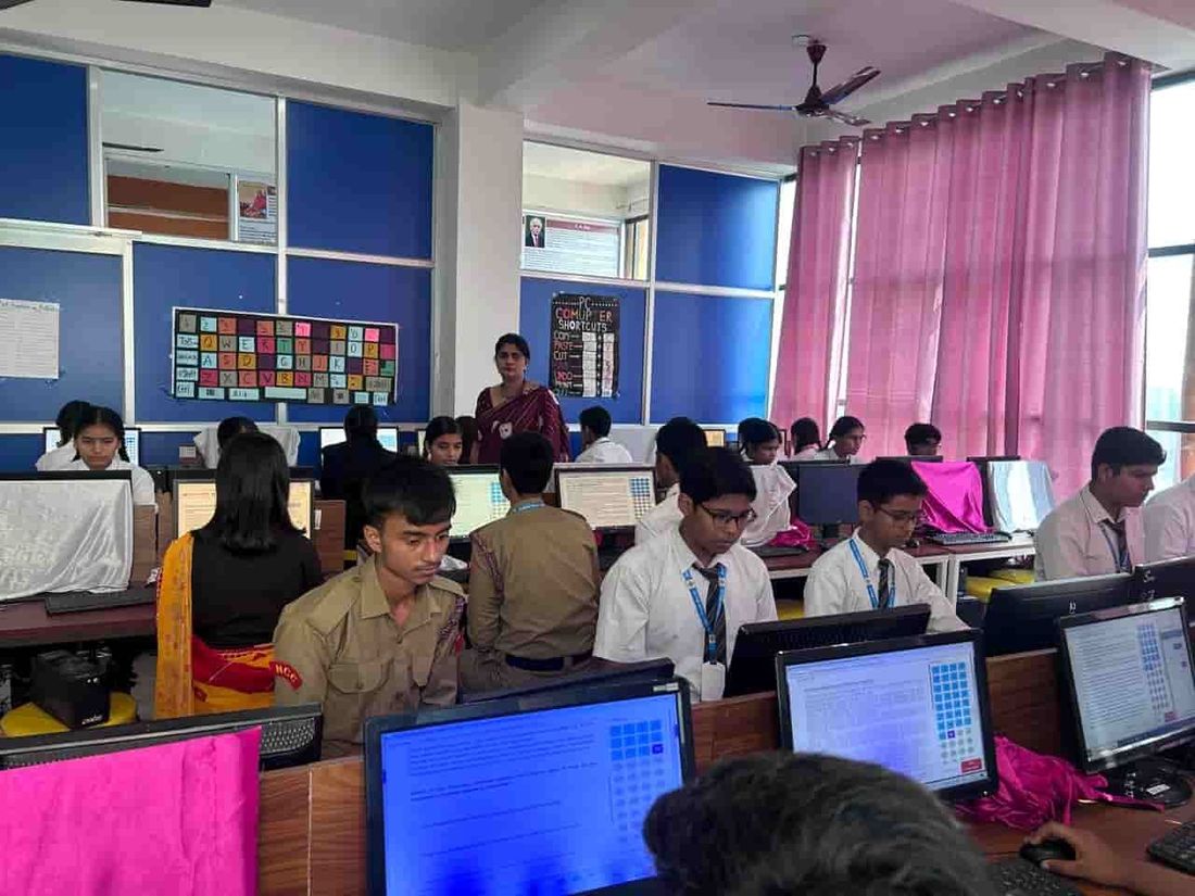 A computer room at Sunbeam School Ballia during a lesson — a teacher at the front and wall charts of keyboard keys and PC shortcuts behind the machines