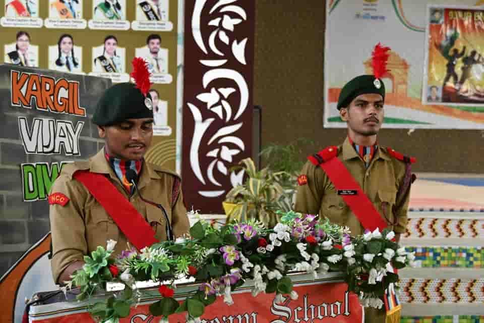 An NCC cadet of Sunbeam School Ballia speaking at the lectern in uniform, a second cadet standing to attention beside him, under a Kargil Vijay Diwas backdrop.