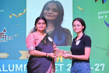 An alumna of Sunbeam School Ballia taking her medallion from a teacher, both holding it between them, her photograph on the screen behind.