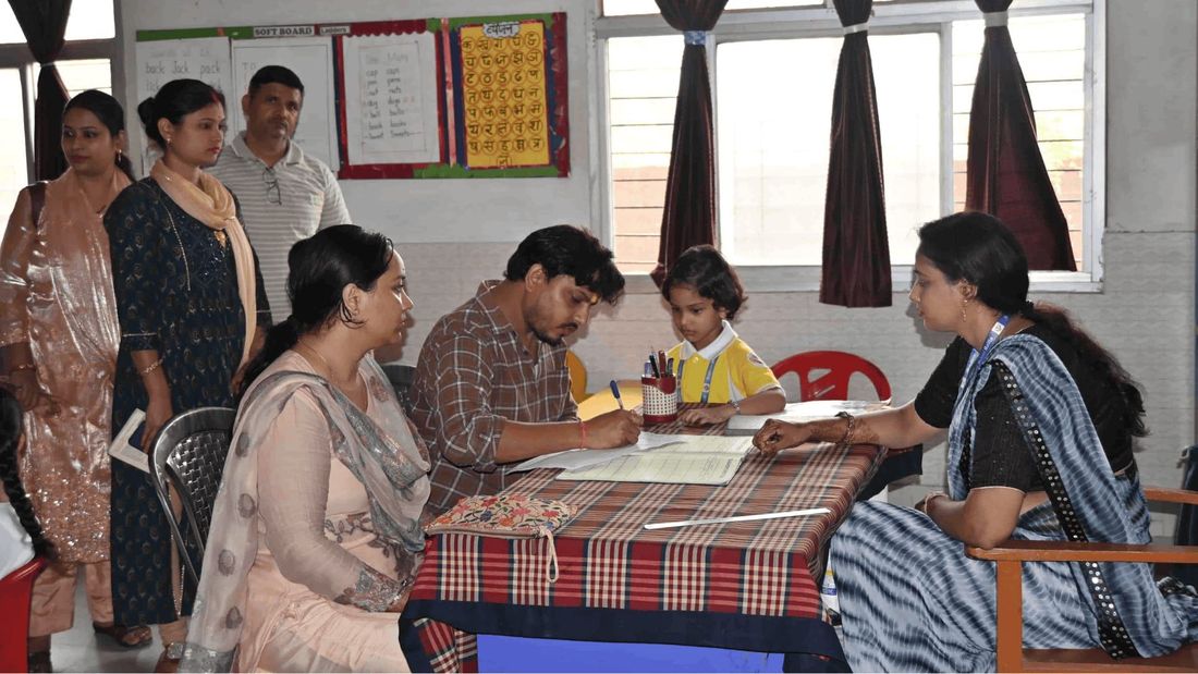 A mother and father at Sunbeam School Ballia completing a form with the class teacher, their child at the desk beside them and other parents waiting their turn