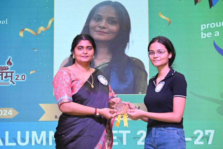 An alumna of Sunbeam School Ballia taking her medallion from a teacher, both holding it between them, her photograph on the screen behind.