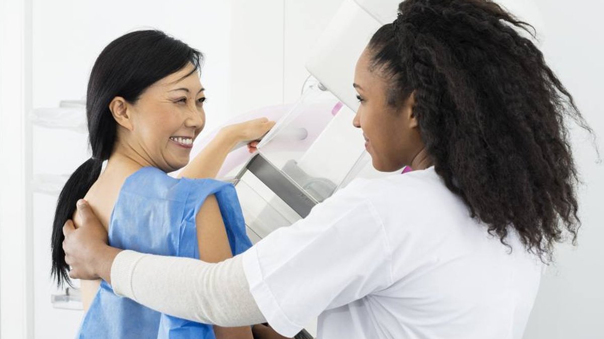 Breast cancer screening becomes more personalized with risk-based guidelines A rear view of a smiling woman undergoing a mammogram. A technician helps assist the woman through the procedure.
