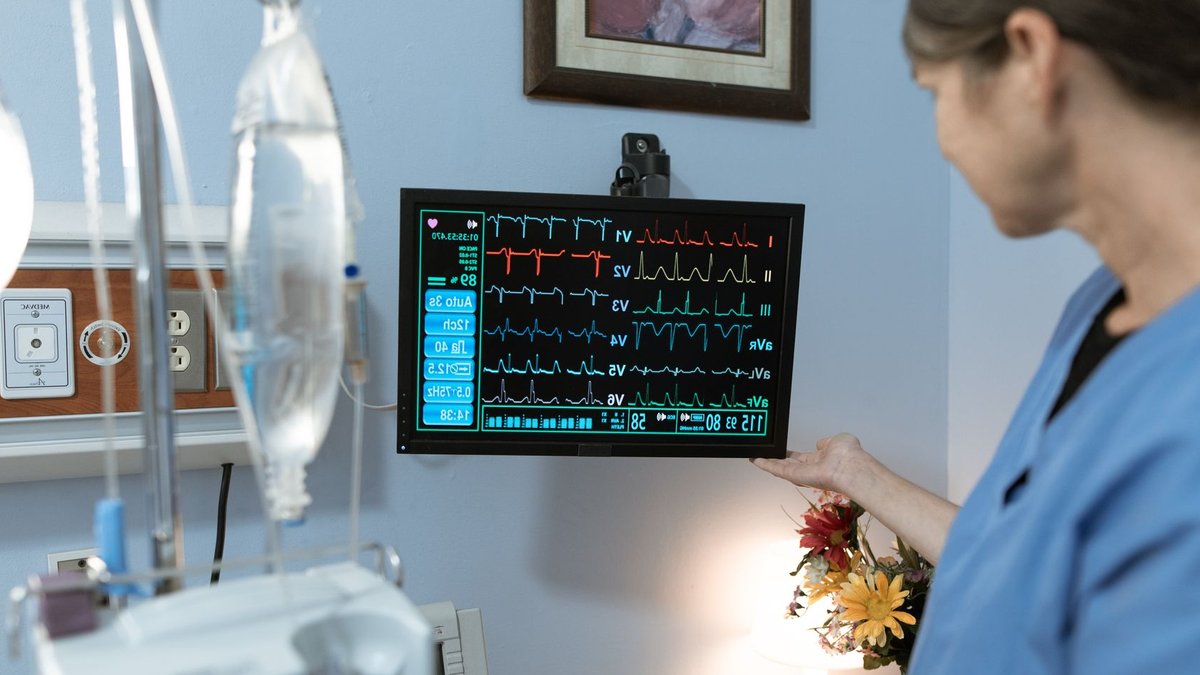 A nurse in blue scrubs examines a medical monitor displaying vital signs in a hospital setting.