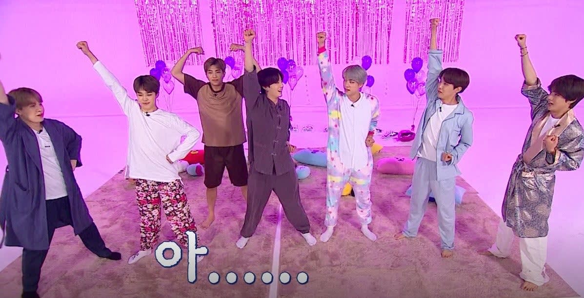 BTS wear pyjamas and have their arms raised on a set decorated with purple streamers and balloons