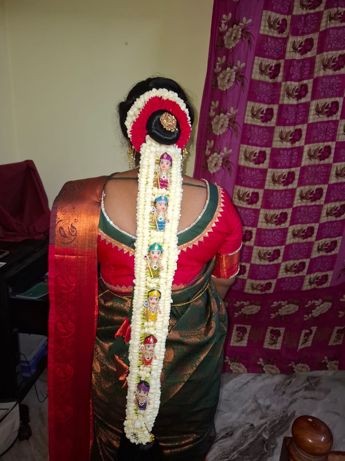 Back view of a bun dressed in white jasmine and red roses, with a pallakku doll garland running the length of the braid over a green and maroon silk saree — bridal services in Chennai