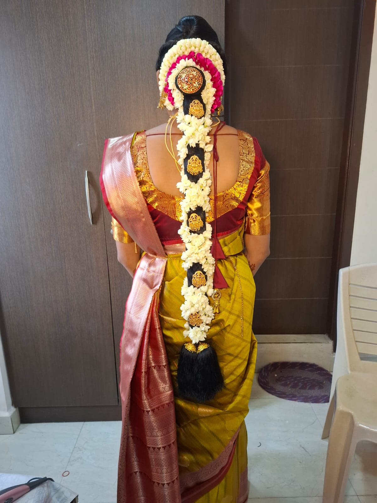 Back view of a bridal braid on a mustard silk saree, decorated with jasmine, pink roses, a jewelled rakodi, gold jadai billai plates and kunjalam tassels — bridal services in Chennai