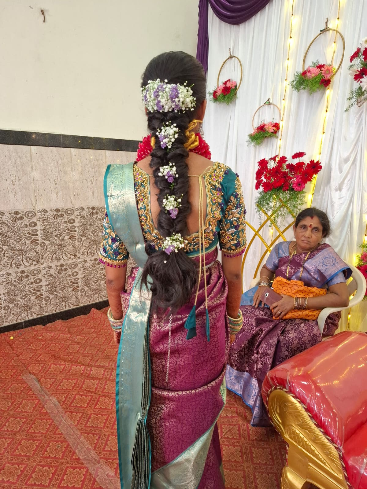 Back view of a fishtail braid threaded with purple and white baby's breath, worn with a magenta silk saree and teal embroidered blouse at a wedding hall — bridal services in Chennai