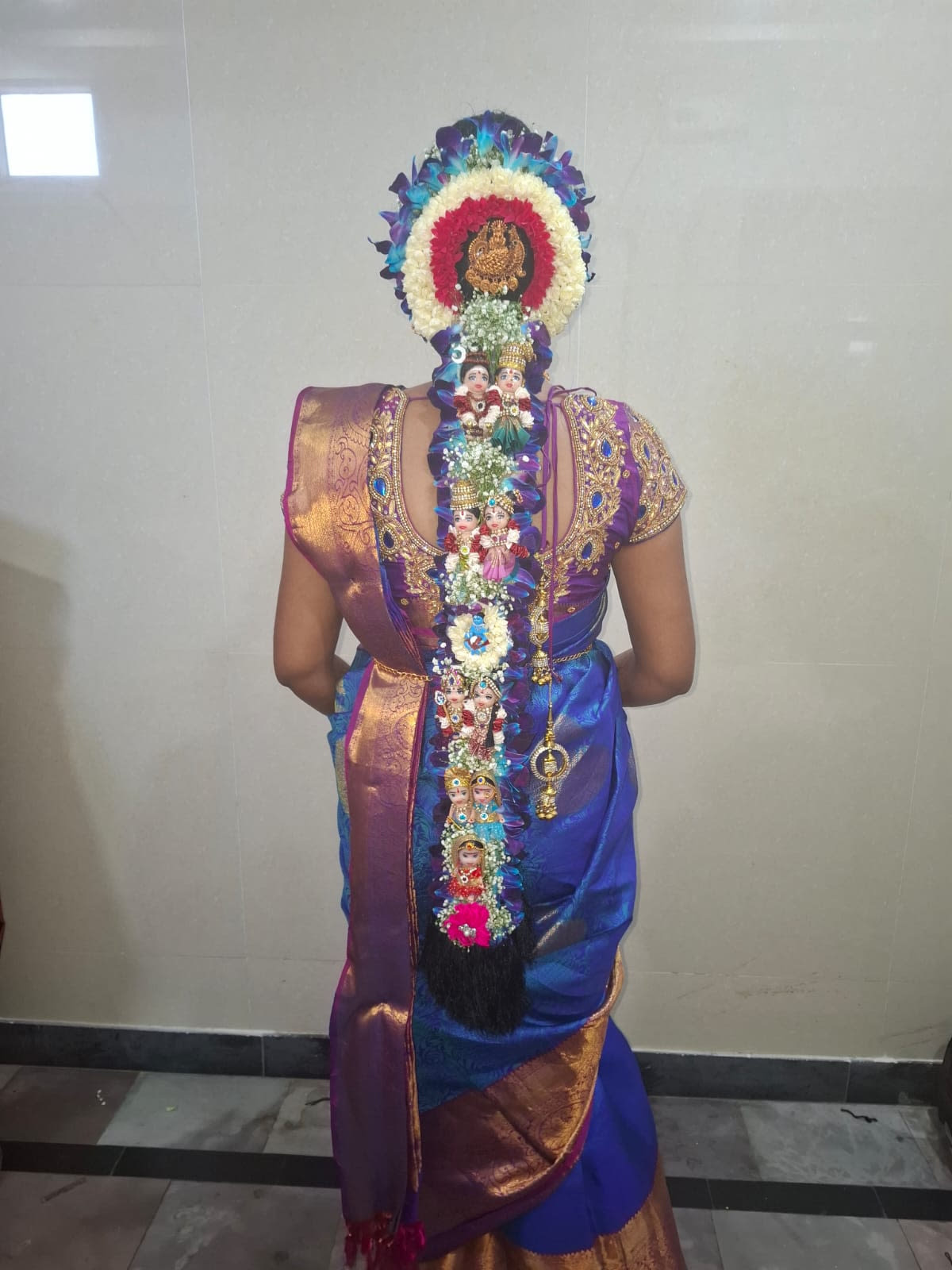Full jadai alangaram on a blue and purple silk saree — blue orchids and jasmine framing a gold Lakshmi pendant, with pallakku dolls and kunjalam down the braid — bridal services in Chennai