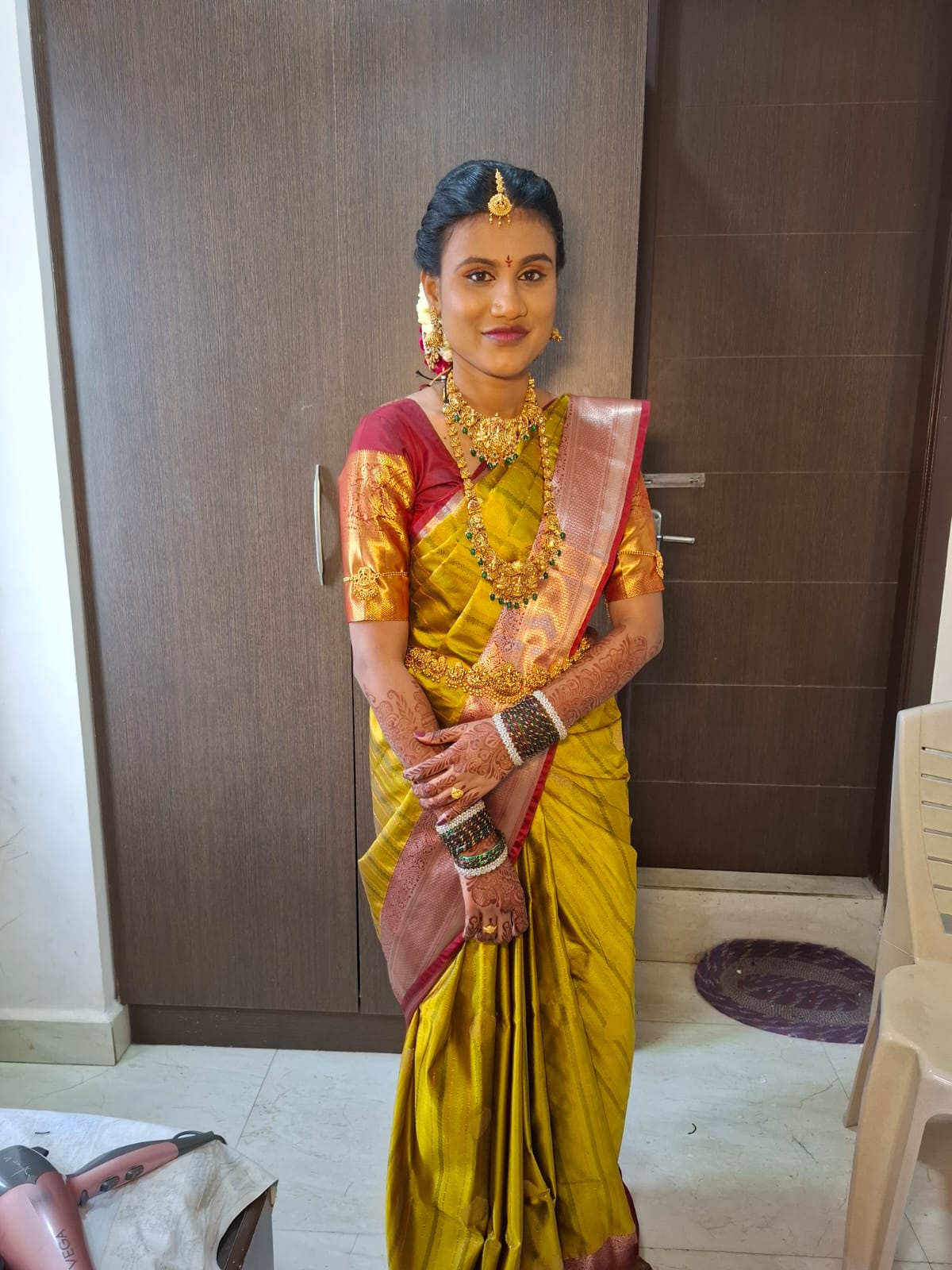 Bride in a mustard silk saree with a red and gold border, Lakshmi temple necklaces, mehendi to the elbow, maang tikka and jasmine at the bun — natural bridal makeup in Chennai
