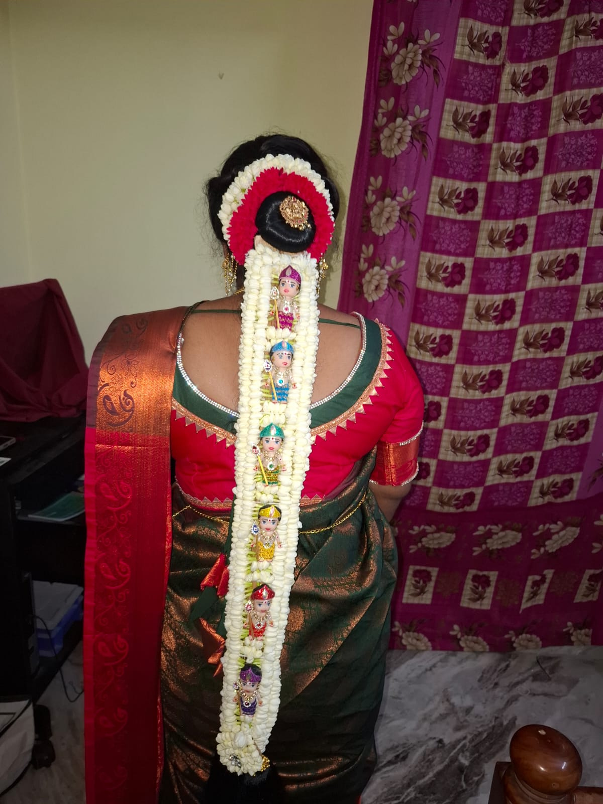 Back view of a bun dressed in white jasmine and red roses, with a pallakku doll garland running the length of the braid over a green and maroon silk saree — madisar saree draping in Chennai