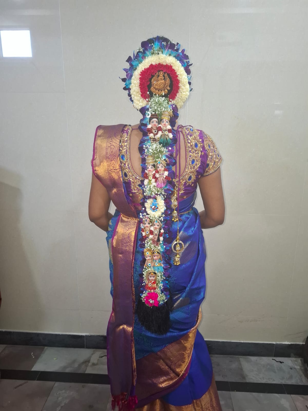 Full jadai alangaram on a blue and purple silk saree — blue orchids and jasmine framing a gold Lakshmi pendant, with pallakku dolls and kunjalam down the braid — South Indian bridal makeup in Chennai