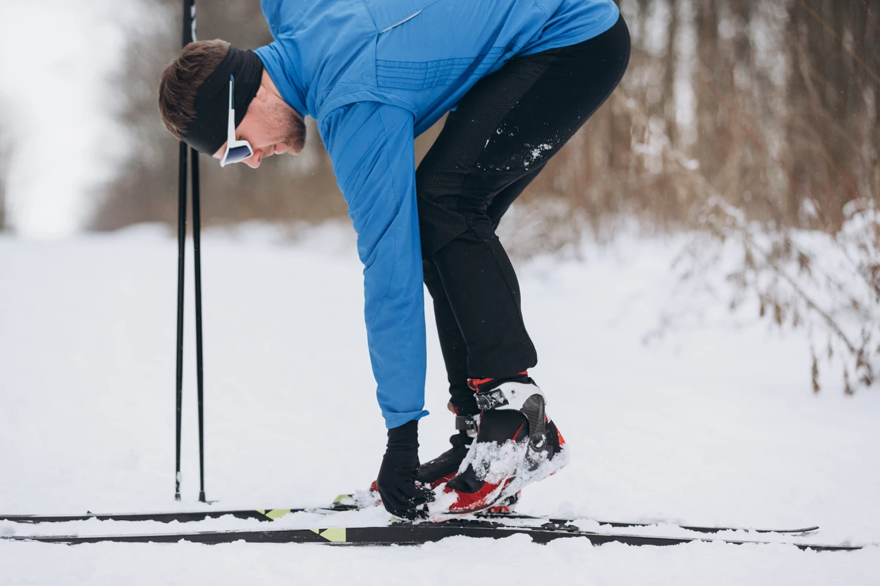 Skieur ajustant la fixation de son ski de fond sur une piste enneigée.