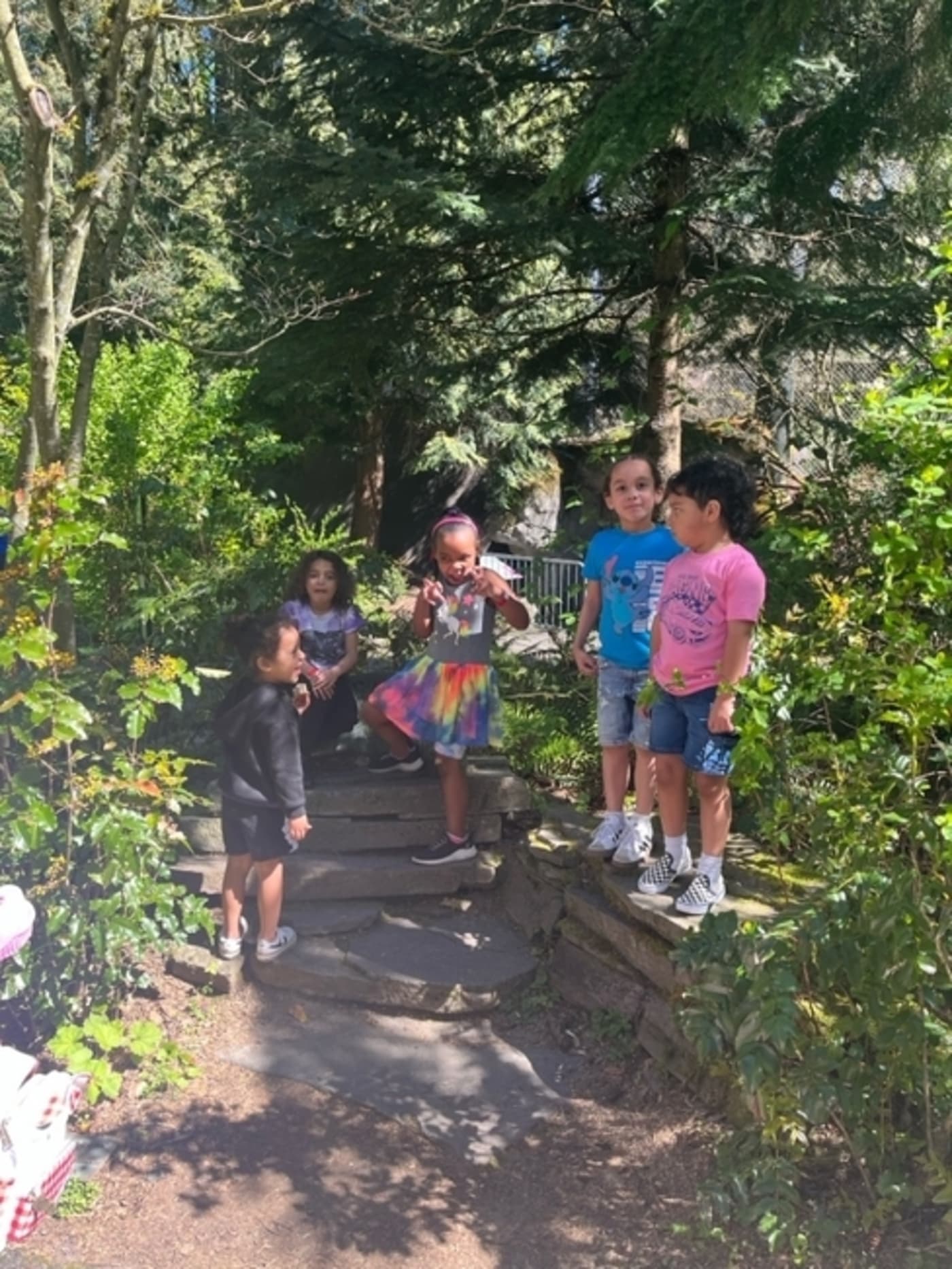  “Group of five young children standing together on a stone path in a lush, green garden, smiling and talking on a sunny day. 