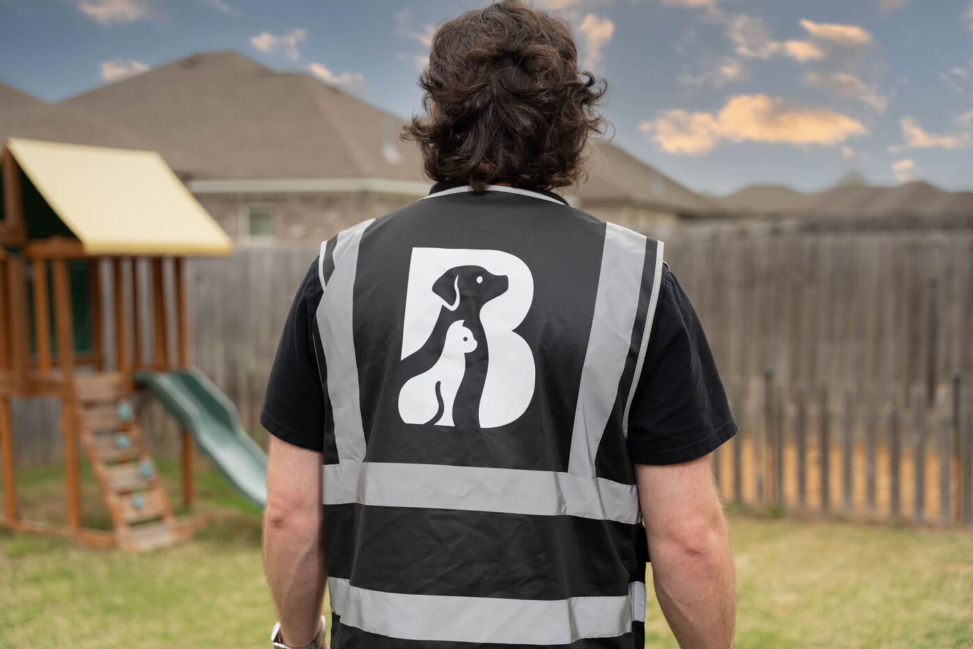 Rear view of a person wearing a black reflective safety vest with a large dog logo on the back, standing in a fenced backyard with a playset in the background at sunset.
