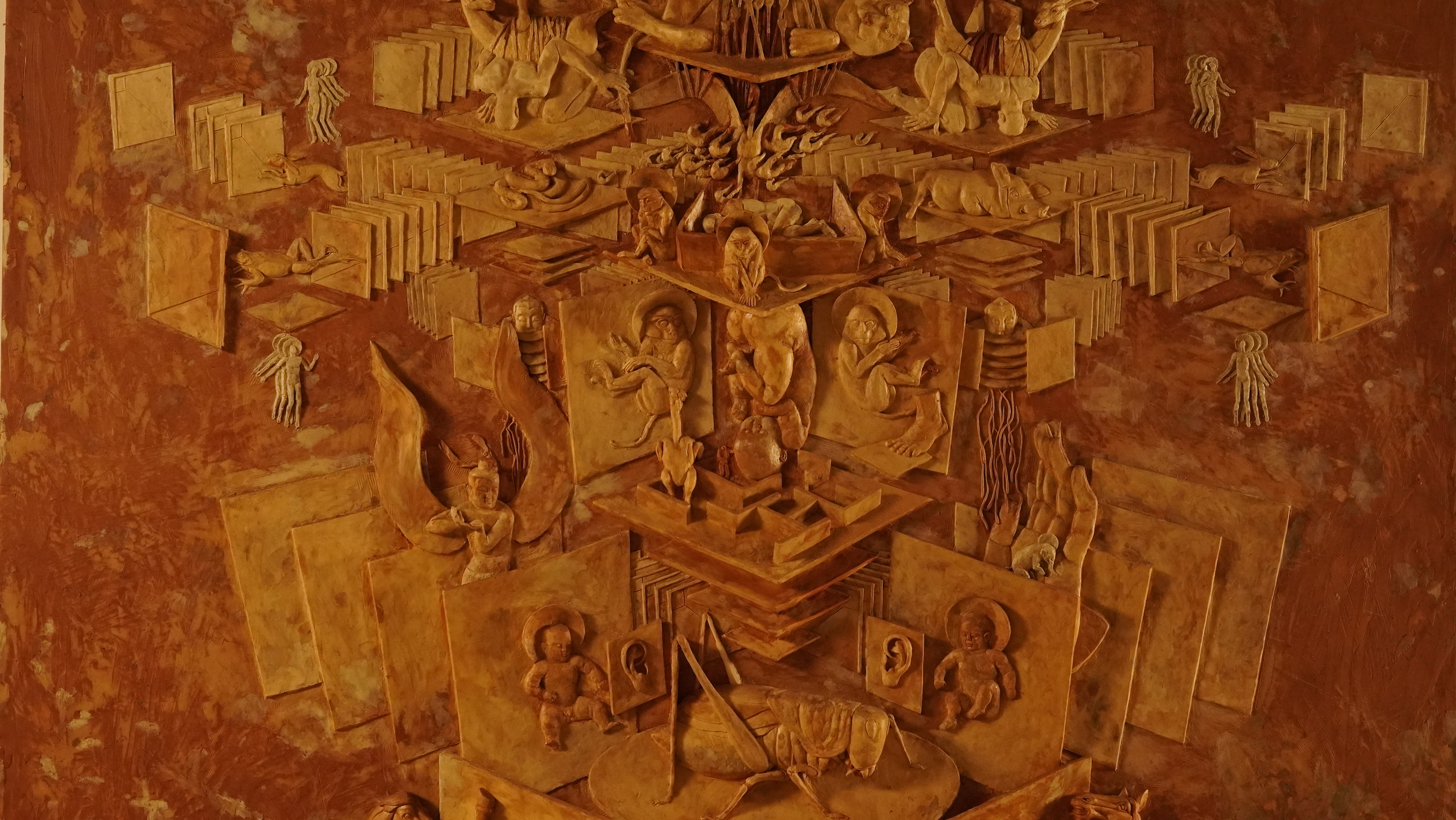Closeup of relief sculpture with many tiny images and shapes: rows of flat square panels moving outward in different directions, monkeys, babies, a large cricket, babies, adult humans curled up like babies, frogs, pigs, snakes, human bent over with deer tied to their backs etc.
