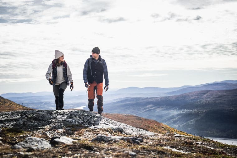 A man and a woman walk towards the camera with a mountainous backdrop wearing outdoor gear including Hestra gloves