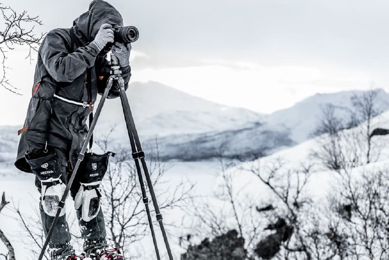 A photographer takes a photo outside with camera mounted on a tripod wearing Hestra glove liners