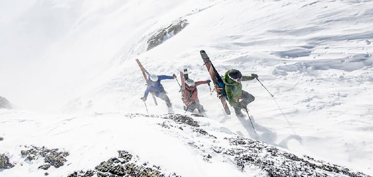 Three skiers bootpack up a steep slope on a windy day