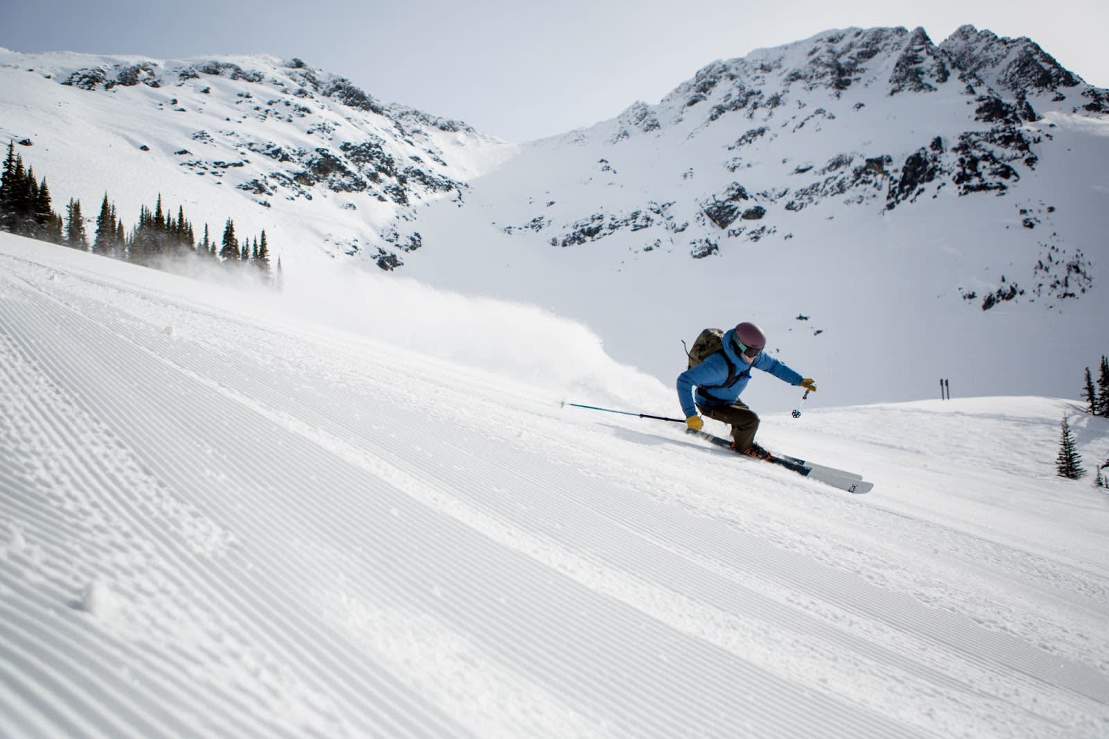 Skier in the foreground on a groomed ski run with snowy mountains in the backdrop