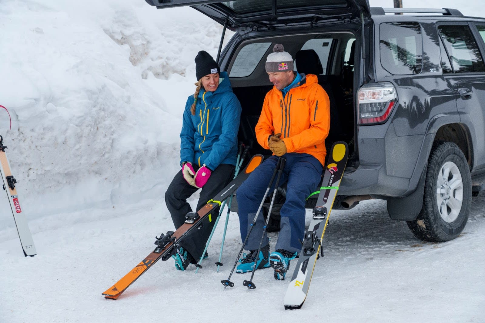 Two skiers sitting on the tailgate of an SUV with their skis and poles