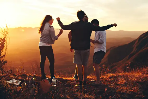 Students stand outside on the top of a hill with the sun shining behind them and mountains in the background