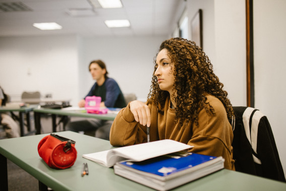 Student sitting at desk in classroom with notebooks, textbook and pencil case; another student out of focus in the background