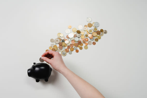 A person places a coin in a black ceramic piggy bank. Other coins are spread out on the table above the bank.