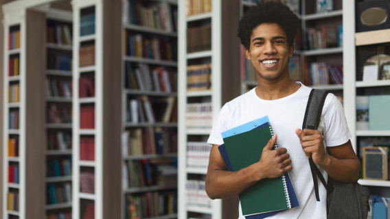 Student smiling, holding notebooks and a backpack in a library