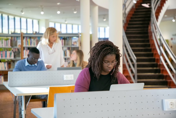 Student working alone in a library, two other students speaking to a professor at the next table back