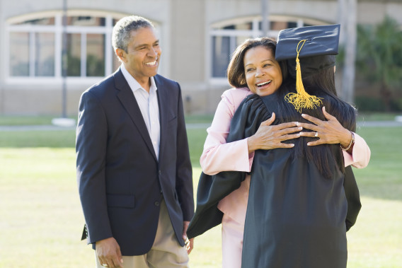 Parent hugging young adult who is wearing a graduation cap and gown, other parent stands close by smiling