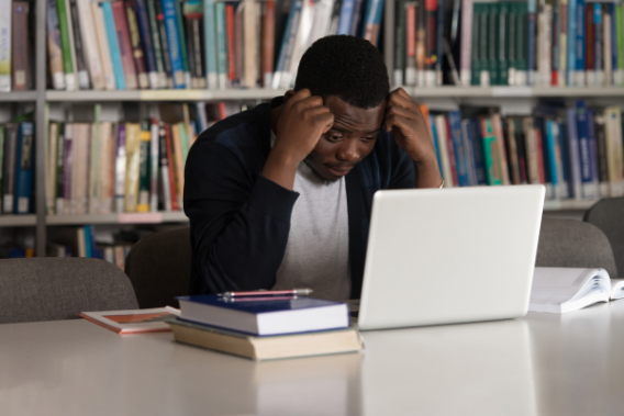 Young man sits at table in library with laptop and a few books, elbows on the table with head resting on hands looking stressed