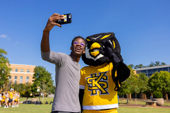 A man holds his phone up to take a picture with an owl mascot